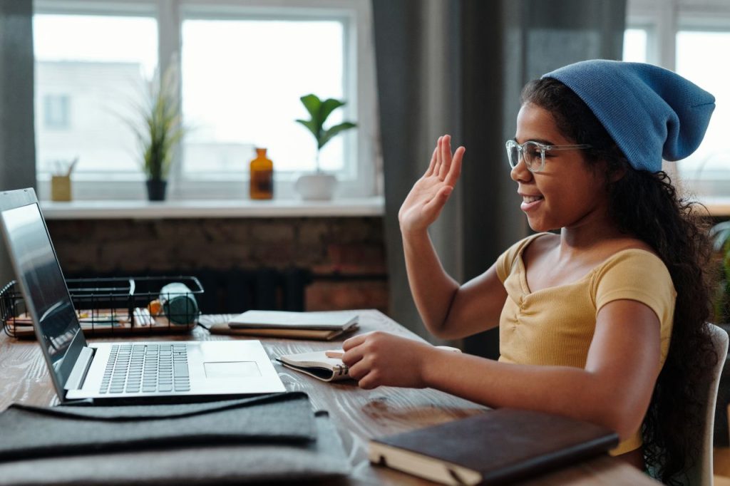 A young girl waves at her laptop during a video call in a cozy home office setting.